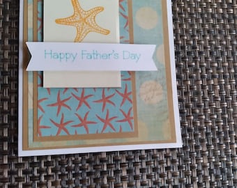 May include: A handmade Father's Day card with a beach theme. The card features a light blue background with a starfish design and the text "Happy Father's Day". The card has a layered design with a beige and white border.