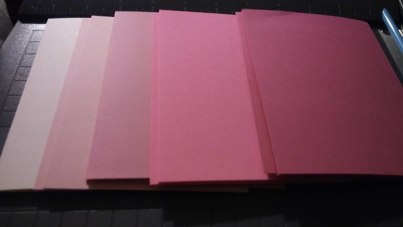 May include: A collection of pink paper sheets in varying shades, from pale blush to deep rose. The papers are neatly stacked, showcasing a gradient of color. The image is well-lit, highlighting the smooth texture of the paper.