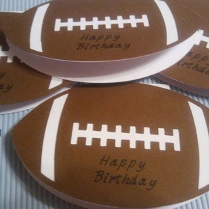Peut inclure: Cartes en forme de ballon de football am&eacute;ricain marron avec des rayures et des coutures blanches. Chaque carte porte l'inscription "Happy Birthday" en noir. Les cartes sont dispos&eacute;es sur une surface &agrave; rayures bleu clair et blanches.