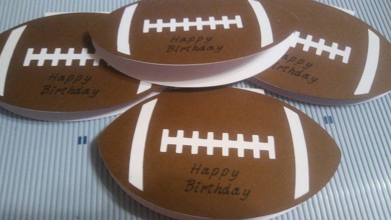 Peut inclure: Cartes en forme de ballon de football am&eacute;ricain marron avec des rayures et des coutures blanches. Chaque carte porte l'inscription "Happy Birthday" en noir. Les cartes sont dispos&eacute;es sur une surface &agrave; rayures bleu clair et blanches.