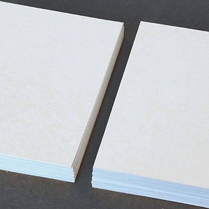 May include: Two stacks of blank, off-white paper. Each stack is rectangular and composed of many individual sheets. The paper has a slightly textured surface. The stacks are positioned side-by-side against a dark background.