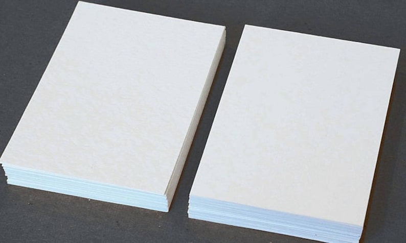 May include: Two stacks of blank, off-white paper. Each stack is rectangular and composed of many individual sheets. The paper has a slightly textured surface. The stacks are positioned side-by-side against a dark background.