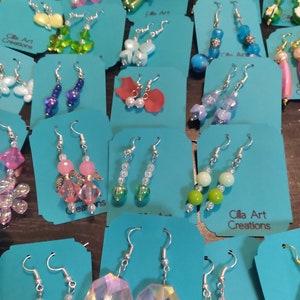 May include: A collection of handmade earrings displayed on teal cards. The earrings feature various designs, including beaded, crystal, and charm styles. Colors include pink, blue, green, and clear. The text "Cilla Art Creations" is visible on some cards.