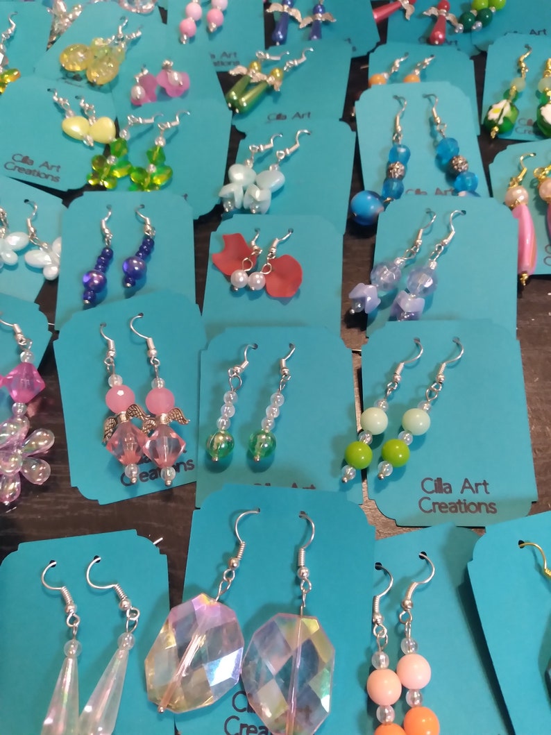 May include: A collection of handmade earrings displayed on teal cards. The earrings feature various designs, including beaded, crystal, and charm styles. Colors include pink, blue, green, and clear. The text "Cilla Art Creations" is visible on some cards.