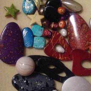 May include: A selection of beads and decorative items in various shapes. Includes star, oval, and irregular forms in colours such as purple, red, black, blue, and white. Some pieces feature speckled or patterned designs.