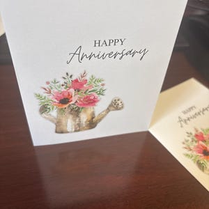 May include: A white greeting card with the words "HAPPY Anniversary" in black script. The card features a watercolor illustration of a watering can overflowing with pink and red flowers and greenery. Another card is partially visible.