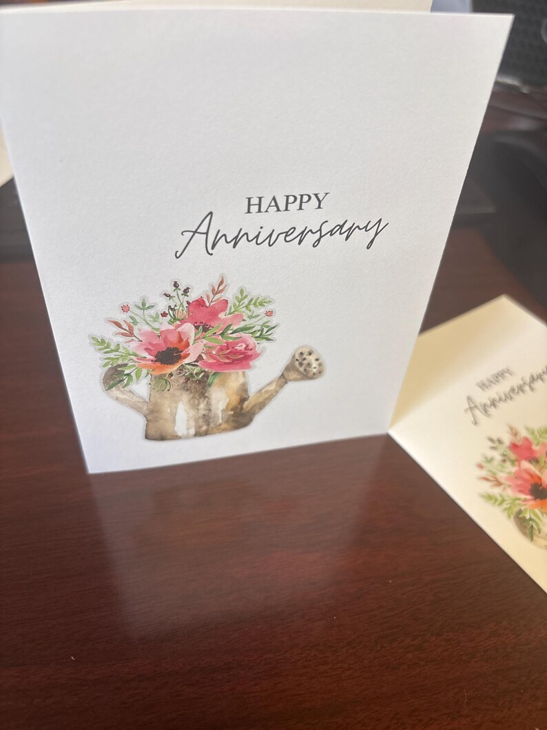 May include: A white greeting card with the words "HAPPY Anniversary" in black script. The card features a watercolor illustration of a watering can overflowing with pink and red flowers and greenery. Another card is partially visible.