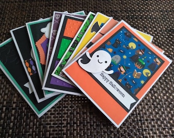 May include: A collection of handmade Halloween greeting cards. The cards feature various designs, including a ghost, witches, bats, and pumpkins. The cards have a variety of colors, including black, orange, green, and purple. The words "Happy Halloween" are printed on a white banner.
