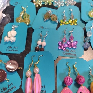 May include: A collection of handmade earrings displayed on teal tags. The earrings feature various designs, including beaded flowers, butterflies, and abstract shapes in colors like pink, yellow, and purple. The text "Cila Art Creations" is visible on some tags.