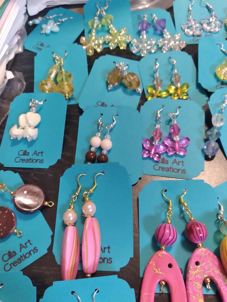 May include: A collection of handmade earrings displayed on teal tags. The earrings feature various designs, including beaded flowers, butterflies, and abstract shapes in colors like pink, yellow, and purple. The text "Cila Art Creations" is visible on some tags.