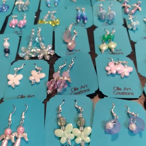 May include: A collection of handmade earrings displayed on teal cards. The earrings feature various designs, including butterflies, flowers, and beaded accents in colors like pink, white, and blue. The text "Cilla Art Creations" is visible on some cards.