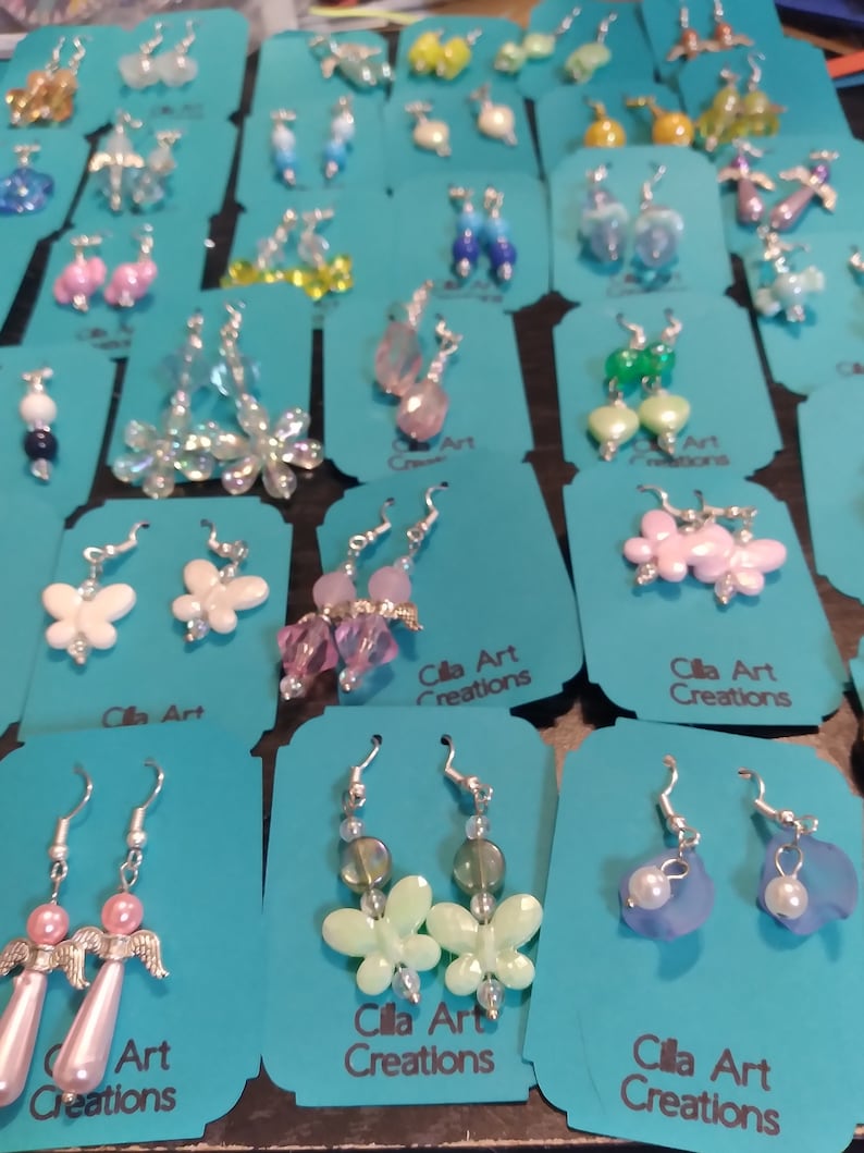 May include: A collection of handmade earrings displayed on teal cards. The earrings feature various designs, including butterflies, flowers, and beaded accents in colors like pink, white, and blue. The text "Cilla Art Creations" is visible on some cards.