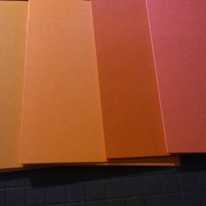 May include: A collection of six rectangular sheets of paper in varying shades of orange, from light peach to deep red-orange. The sheets are stacked, with the edges visible, creating a gradient effect. The paper appears to be smooth and matte.