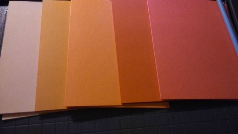 May include: A collection of six rectangular sheets of paper in varying shades of orange, from light peach to deep red-orange. The sheets are stacked, with the edges visible, creating a gradient effect. The paper appears to be smooth and matte.