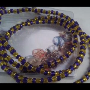 May include: A beaded necklace featuring alternating gold and purple beads. The necklace is made of small, round beads strung together to create a layered design. The necklace is displayed on a white surface.
