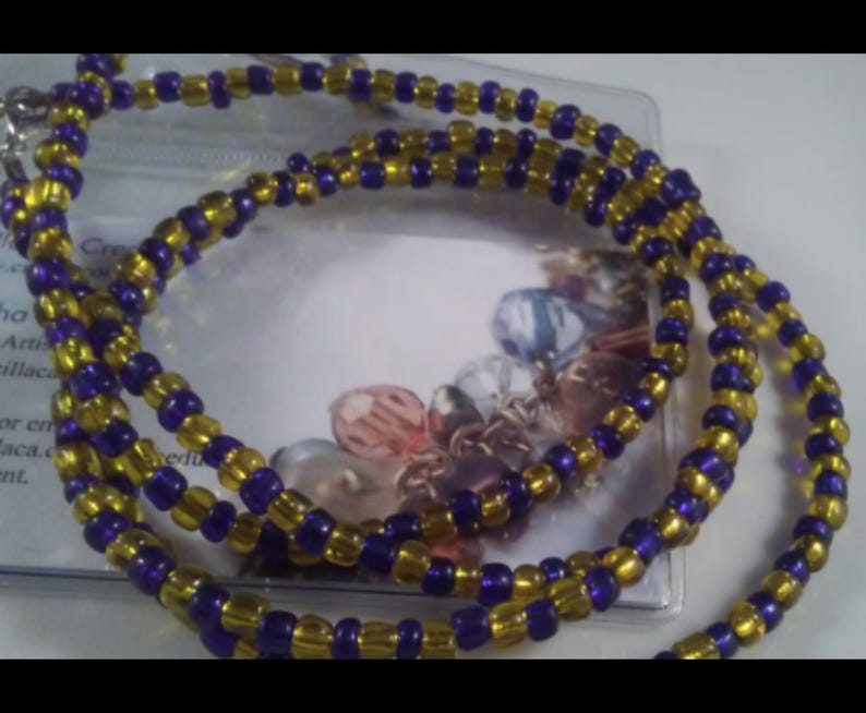 May include: A beaded necklace featuring alternating gold and purple beads. The necklace is made of small, round beads strung together to create a layered design. The necklace is displayed on a white surface.