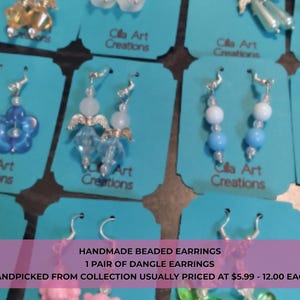 May include: A collection of handmade beaded dangle earrings displayed on teal cards. The earrings feature various designs, including flower shapes, angel figures, and beaded strands in shades of blue, white, and clear. Each pair is unique.