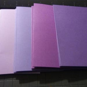 May include: A stack of rectangular paper sheets in shades of purple, from light lavender to deep violet. The papers are neatly arranged, with a blue paper cutter visible in the background. The papers are likely for crafting or stationery.