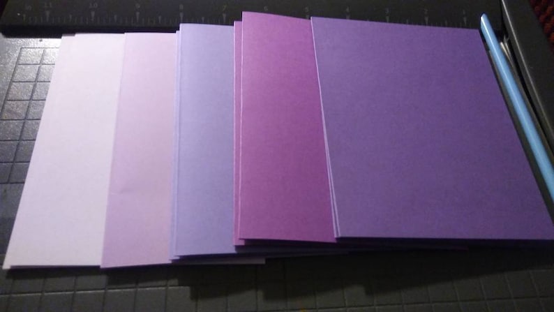 May include: A stack of rectangular paper sheets in shades of purple, from light lavender to deep violet. The papers are neatly arranged, with a blue paper cutter visible in the background. The papers are likely for crafting or stationery.
