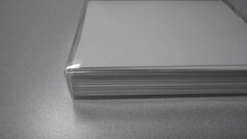 Op de afbeelding: Een stapel witte papieren vellen in een doorzichtige plastic houder. Het papier is netjes opgestapeld en de plastic houder is transparant. De afbeelding is in grijstinten, met de nadruk op het papier en de beschermende hoes.
