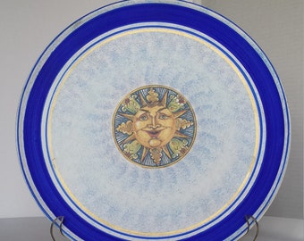 May include: A blue and white ceramic plate with a gold rim and a painted sun face in the centre. The sun has a yellow face with a smile and closed eyes, and is surrounded by gold rays and green leaves.