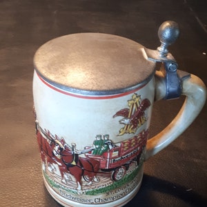 K&ouml;nnte beinhalten: Ein Budweiser Bierkrug aus Keramik mit Deckel und Henkel. Der Krug zeigt einen roten Wagen, der von zwei Pferden gezogen wird, mit einem Budweiser-Logo und dem Text "Budweiser, Champion of Beers, St. Louis, Mo."