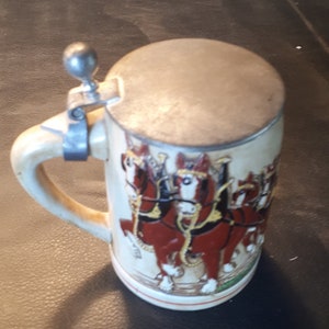 K&ouml;nnte beinhalten: Ein wei&szlig;er Keramik-Bierkrug mit einem silbernen Deckel und einem Henkel. Der Krug zeigt ein farbenfrohes Bild von sechs braunen Pferden, die einen Wagen ziehen.