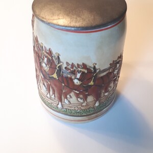 K&ouml;nnte beinhalten: Ein Keramik-Bierkrug mit einem Zinn-Deckel und einer Darstellung eines Gespanns mit acht braunen Pferden, die einen Wagen ziehen. Der Krug ist mit gr&uuml;nen, roten und wei&szlig;en Akzenten verziert.
