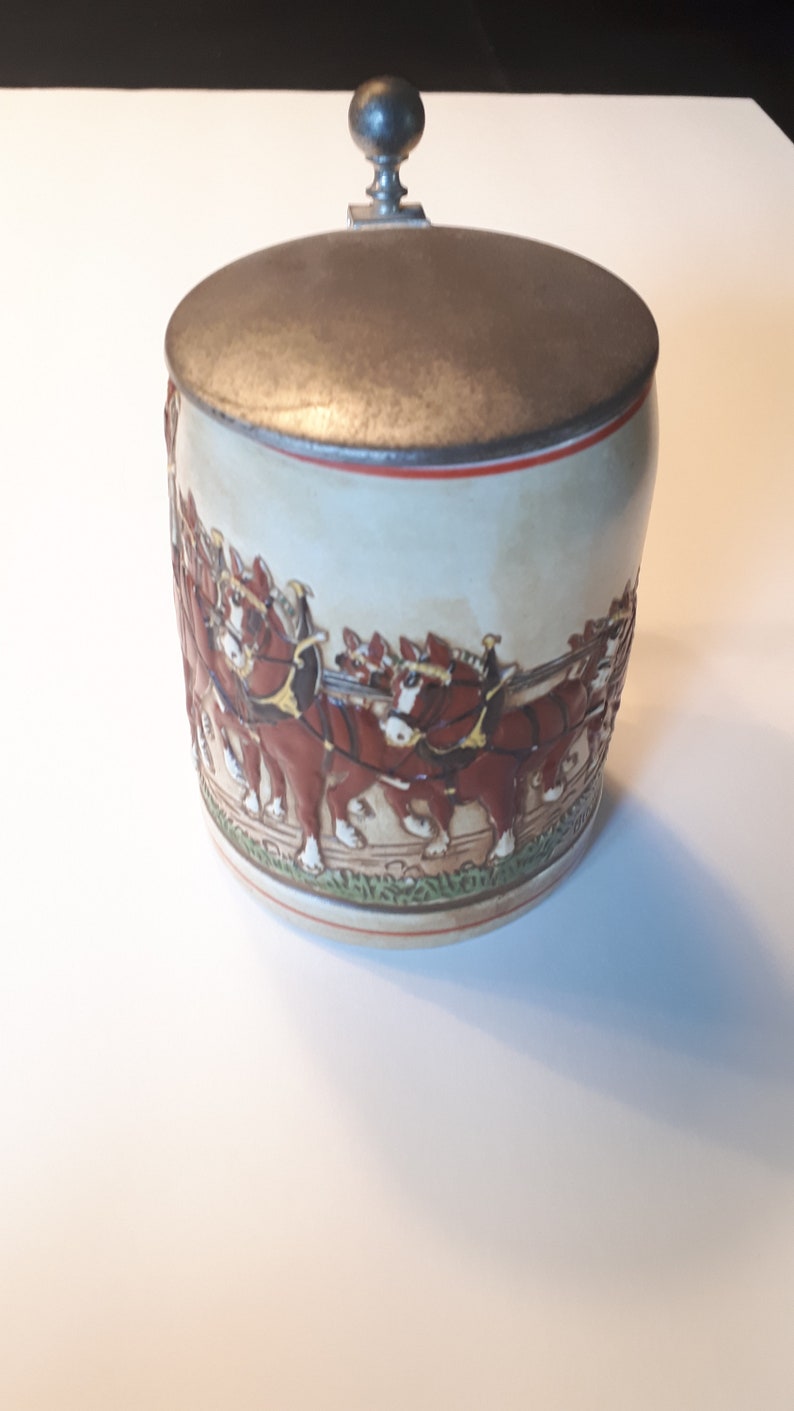K&ouml;nnte beinhalten: Ein Keramik-Bierkrug mit einem Zinn-Deckel und einer Darstellung eines Gespanns mit acht braunen Pferden, die einen Wagen ziehen. Der Krug ist mit gr&uuml;nen, roten und wei&szlig;en Akzenten verziert.