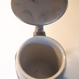 K&ouml;nnte beinhalten: Ein Bierkrug aus Keramik mit Deckel. Der Krug hat ein braunes und wei&szlig;es Design mit einem Blumenmuster. Der Deckel ist aus Metall und hat einen Griff.
