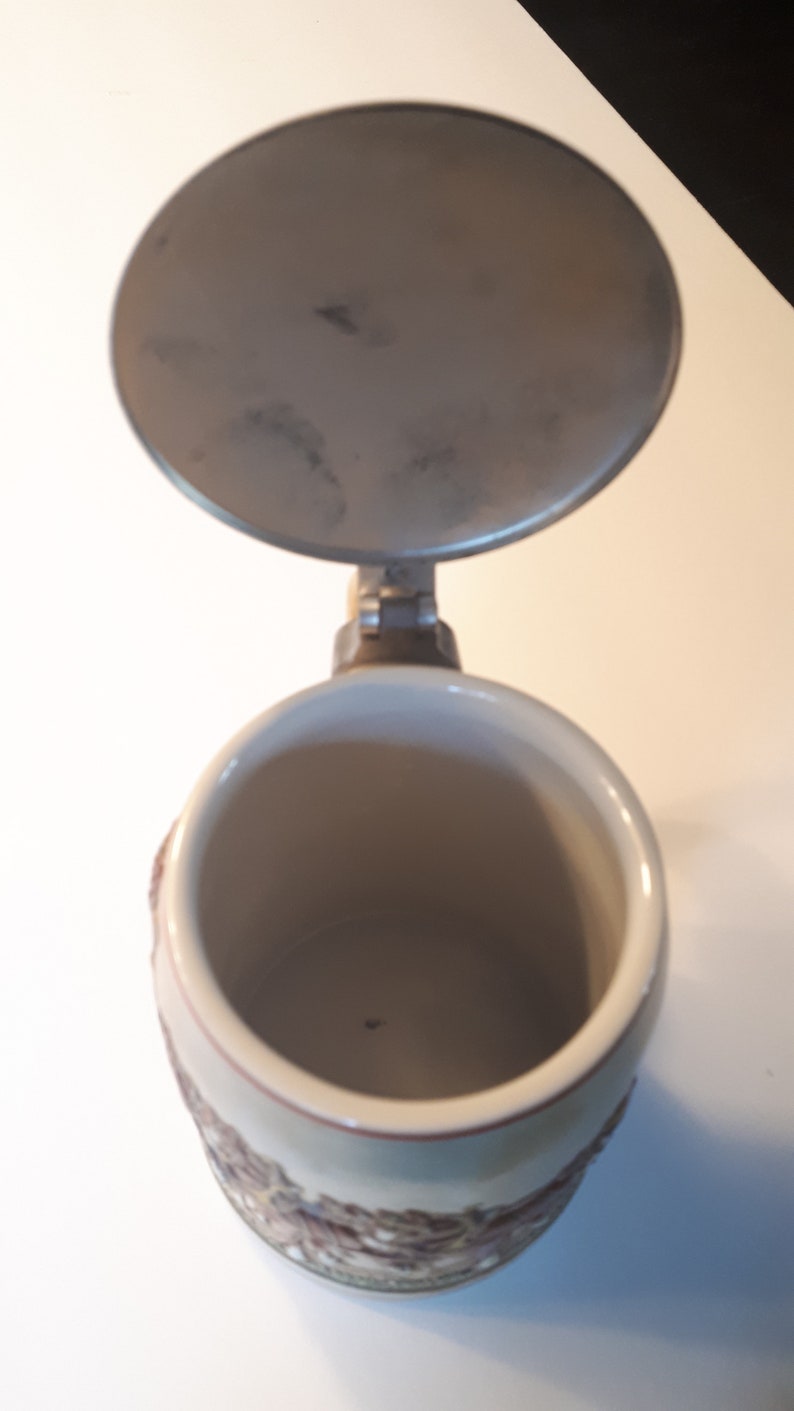 K&ouml;nnte beinhalten: Ein Bierkrug aus Keramik mit Deckel. Der Krug hat ein braunes und wei&szlig;es Design mit einem Blumenmuster. Der Deckel ist aus Metall und hat einen Griff.