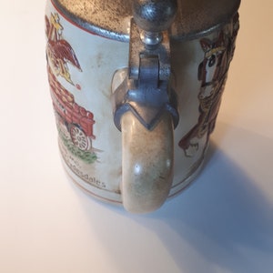 K&ouml;nnte beinhalten: Ein antiker Bierkrug aus Keramik mit einem Zinnverschluss und -griff. Der Krug zeigt eine farbenfrohe Illustration eines von Pferden gezogenen Wagens und den Text "And desdales".