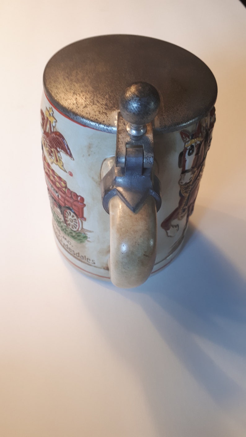 K&ouml;nnte beinhalten: Ein antiker Bierkrug aus Keramik mit einem Zinnverschluss und -griff. Der Krug zeigt eine farbenfrohe Illustration eines von Pferden gezogenen Wagens und den Text "And desdales".