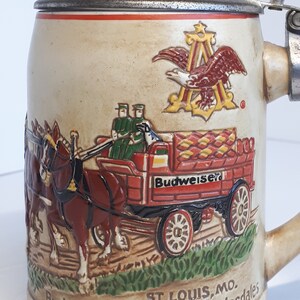 K&ouml;nnte beinhalten: Ein Budweiser Bierkrug aus Keramik mit einem Zinnverschluss und einem Henkel. Der Krug zeigt eine Szene der Budweiser Clydesdales, die einen roten Wagen mit dem Budweiser-Logo ziehen. Der Krug zeigt auch das Budweiser-Logo und den Text "Budweiser, Champion Clydesdales, St. Louis, Mo."