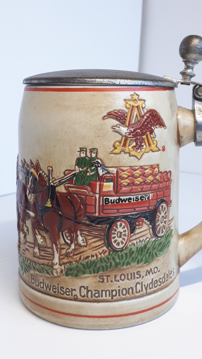 K&ouml;nnte beinhalten: Ein Budweiser Bierkrug aus Keramik mit einem Zinnverschluss und einem Henkel. Der Krug zeigt eine Szene der Budweiser Clydesdales, die einen roten Wagen mit dem Budweiser-Logo ziehen. Der Krug zeigt auch das Budweiser-Logo und den Text "Budweiser, Champion Clydesdales, St. Louis, Mo."