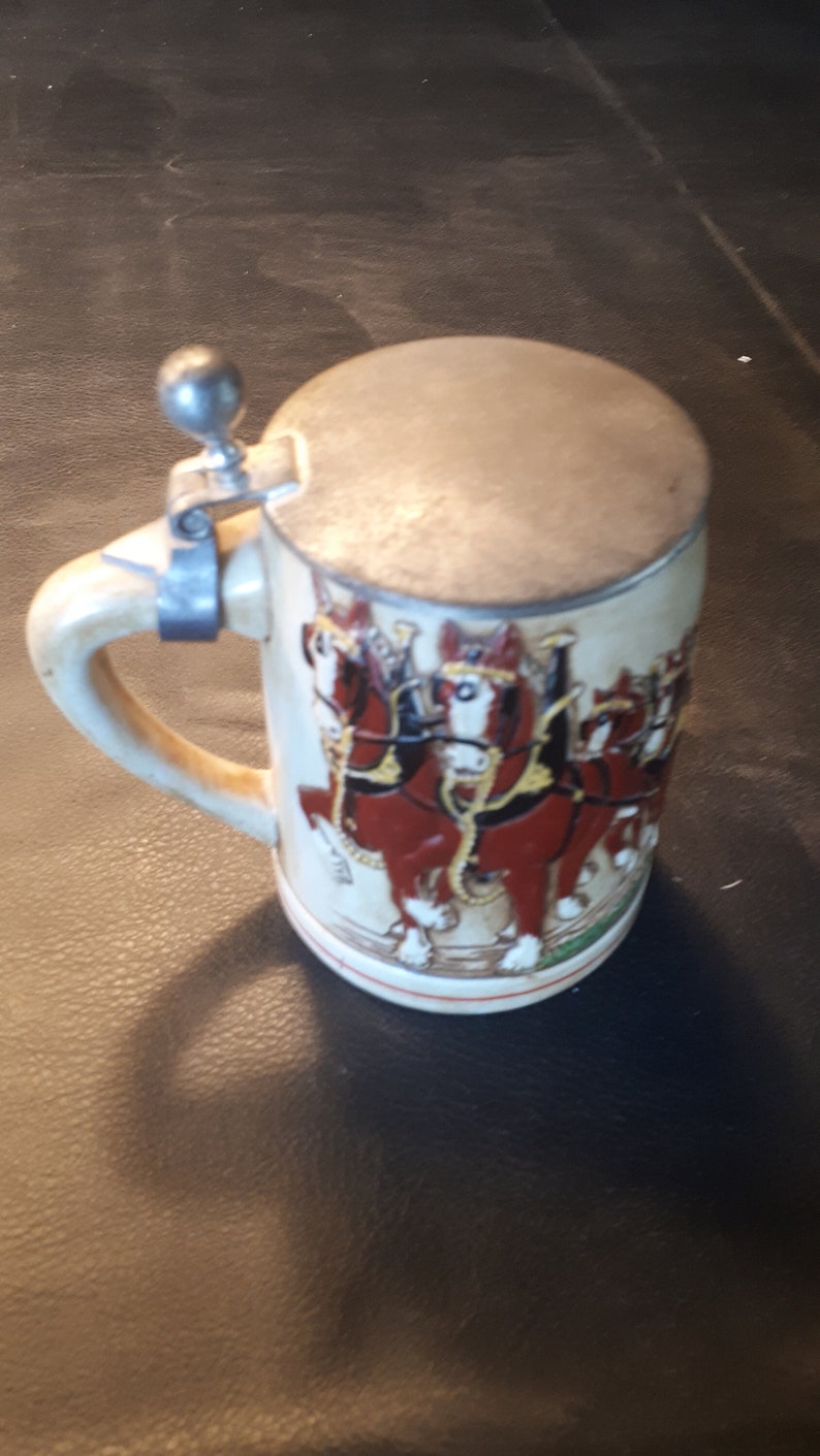 K&ouml;nnte beinhalten: Ein wei&szlig;er Keramik-Bierkrug mit einem silbernen Deckel und einem Henkel. Der Krug zeigt ein farbenfrohes Bild von sechs braunen Pferden, die einen Wagen ziehen.