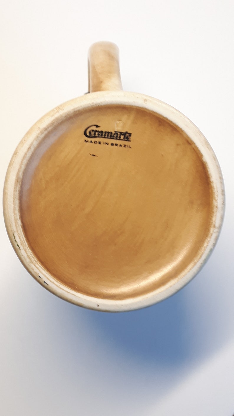 K&ouml;nnte beinhalten: Die Unterseite einer beigefarbenen Keramiktasse mit braunem Innenraum. Die Tasse hat einen schwarzen Stempel mit der Aufschrift "Ceramarte Made in Brazil".