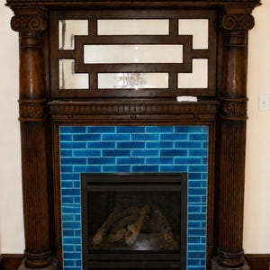 May include: A dark wood fireplace with a blue tile surround. The fireplace features a decorative wooden mantel with a mirrored panel and two ornate columns. The firebox has a black screen and contains faux logs.