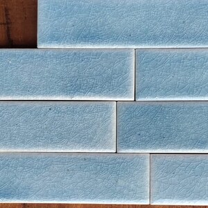 May include: A close-up of four light blue ceramic tiles with a crackled glaze finish. The tiles are arranged in a staggered pattern.