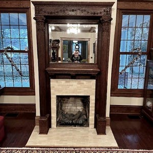 May include: A dark wood fireplace mantel with a rectangular mirror, flanked by two windows. The fireplace has a brick surround and a firebox with logs. The room has a rug with a floral pattern.