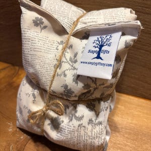 May include: A stack of two fabric pouches tied with twine. The pouches are cream-colored with a gray floral and newspaper print. A white tag with a tree logo and the text "Simply Gifts" is attached.