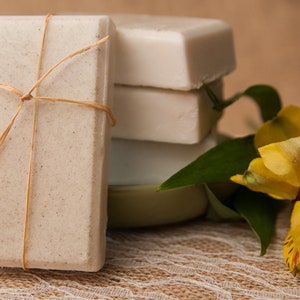 May include: A stack of handmade soaps, one wrapped with twine, next to a bouquet of yellow flowers. The soaps are various shades of white and beige, suggesting natural ingredients. The image evokes a sense of organic beauty and self-care.