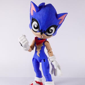 Puede incluir: Una figura de acción azul y amarilla con un pañuelo rojo y guantes blancos. La figura tiene un aspecto estilizado y de dibujos animados con ojos grandes y una amplia sonrisa. La figura está en pose con un brazo levantado y un puño cerrado.