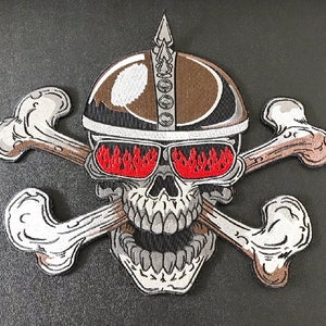 Könnte beinhalten: Gestickter Aufnäher mit einem weißen Totenkopf, der einen braunen Helm mit einem silbernen Dorn auf der Oberseite trägt. Der Schädel hat rote Augen mit Flammen und hält gekreuzte weiße Knochen.