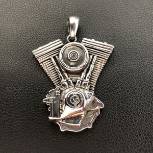 Può includere: Un ciondolo d'argento a forma di motore di moto dettagliato. Il ciondolo ha un anello nella parte superiore per attaccarlo a una catena.