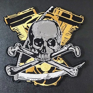 Könnte beinhalten: Ein gestickter Patch mit einem silbernen Totenkopf und gekreuzten Knochen auf schwarzem Hintergrund und einem goldenen Motorrad-Motoren-Design.