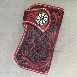 Könnte beinhalten: Rote und schwarze Ledergeldbörse mit floralem und scrollendem Design. Die Geldbörse hat einen dekorativen Silberknopf mit einem Totenkopf-Emblem. Weiße Nähte umranden die Kanten der Geldbörse.