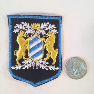 May include: A black embroidered patch with a blue border, featuring a coat of arms with two golden lions flanking a blue and white shield. The shield has a white crown on top and is surrounded by white floral designs.