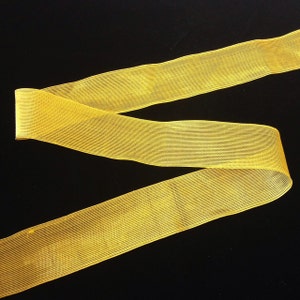 May include: A yellow, sheer ribbon with a textured, woven pattern. The ribbon is folded in half and laid flat on a black surface.