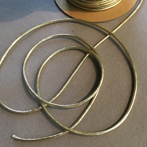 May include: Gold metallic cord, a type of trim or embellishment, is coiled on a surface. The cord has a textured, snake-like appearance.